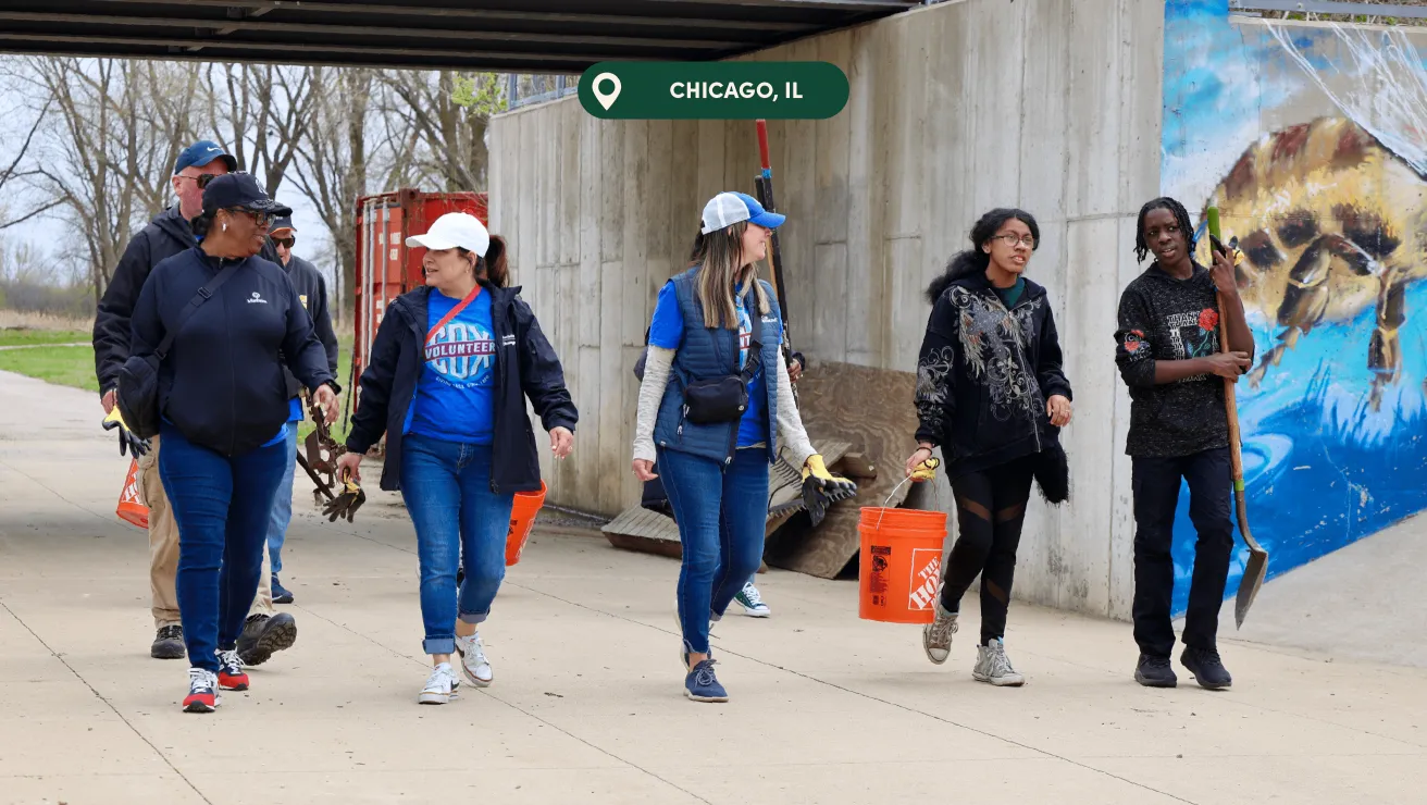 A group of volunteers walks along a pathway in Chicago, carrying tools and buckets, with a colorful mural on the wall beside them.