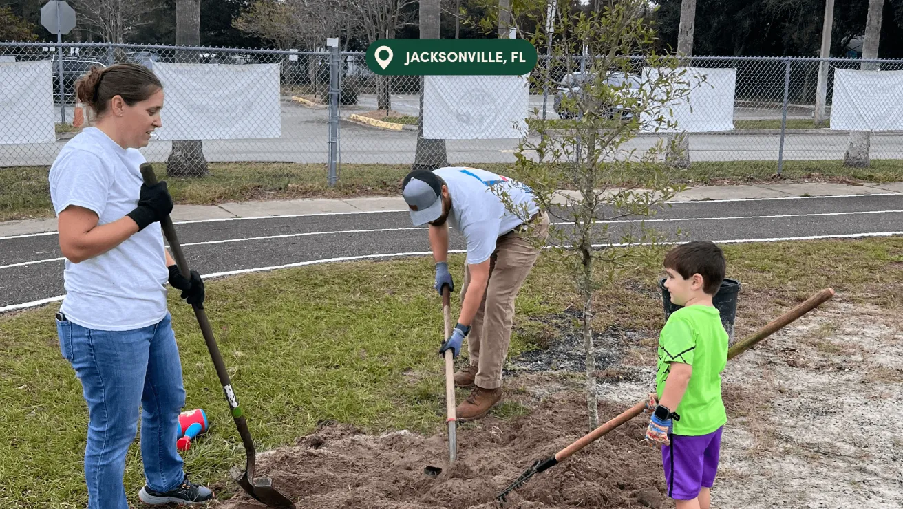 Three individuals participate in a community tree planting event in Jacksonville, FL, surrounded by grass and a chain-link fence.