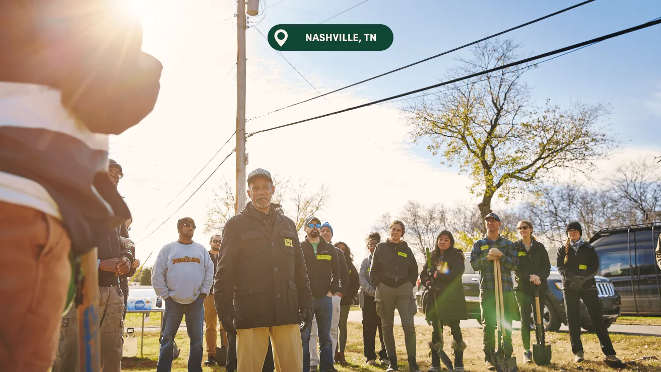 A diverse group of individuals gather outdoors in Nashville, TN, facing an organizer while holding shovels under a bright, sunny sky.