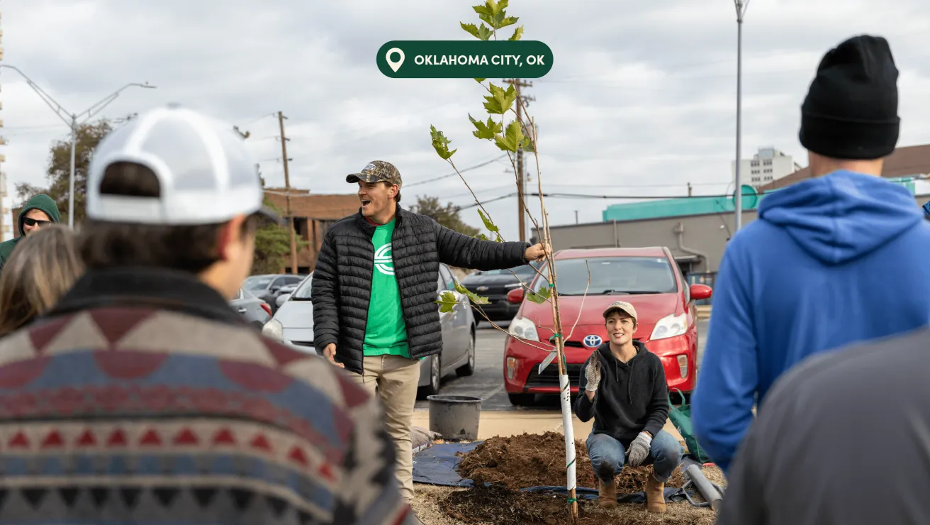 A group gathers in Oklahoma City for a tree planting demonstration, featuring a speaker with a sapling and participants engaged in discussion.