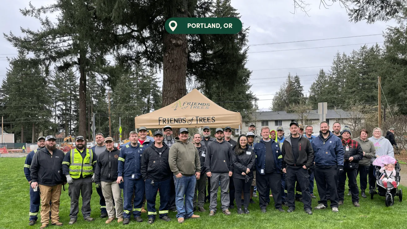 A diverse group of volunteers stands in front of a Friends of Trees tent in a park in Portland, OR, surrounded by tall evergreens.