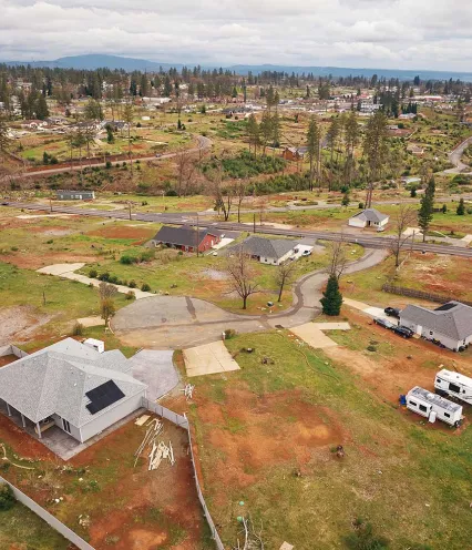Aerial view of a suburban area with scattered houses and open land, surrounded by trees under a cloudy sky.