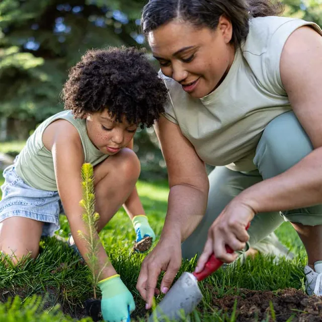 A woman and a child are gardening together, digging in lush green grass with small gardening tools and wearing gloves.