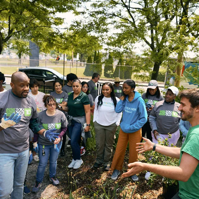 A diverse group of people gathers outdoors for a gardening activity, wearing casual clothing. The atmosphere is collaborative and lively, with trees in the background.