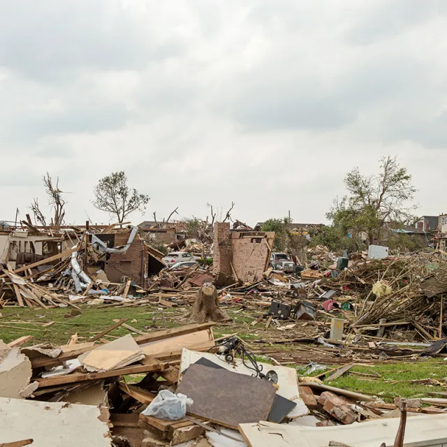 Debris and rubble covering a large area after a tornado, with remains of destroyed homes and vehicles. Overcast sky adds to the somber mood.
