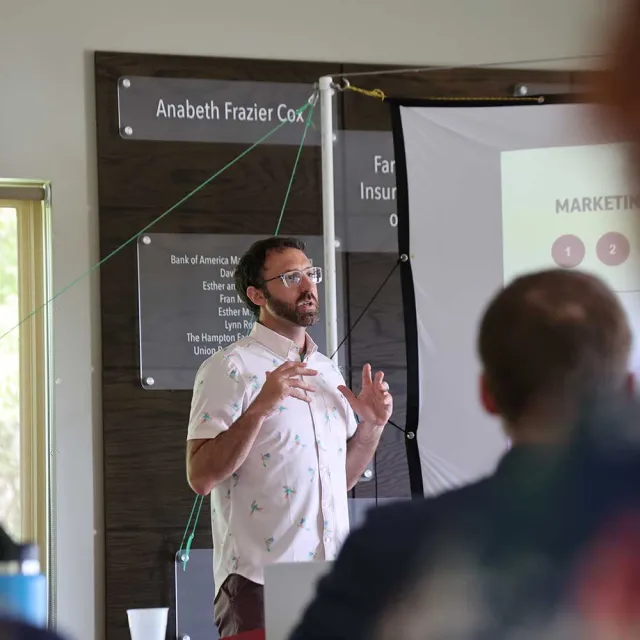 A speaker stands in front of a projector screen during a presentation, gesturing with enthusiasm to an audience in a conference room.
