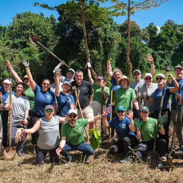 A group of enthusiastic volunteers poses outdoors, holding shovels and planting tools, surrounded by lush greenery and a clear blue sky.