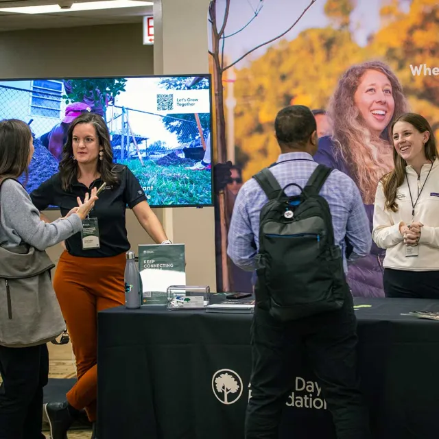 A group of people discuss initiatives at a booth featuring promotional materials and a large display screen about community engagement.