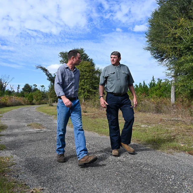 Two men walk along a gravel path in a wooded area, discussing amidst a backdrop of trees and a partly cloudy sky.