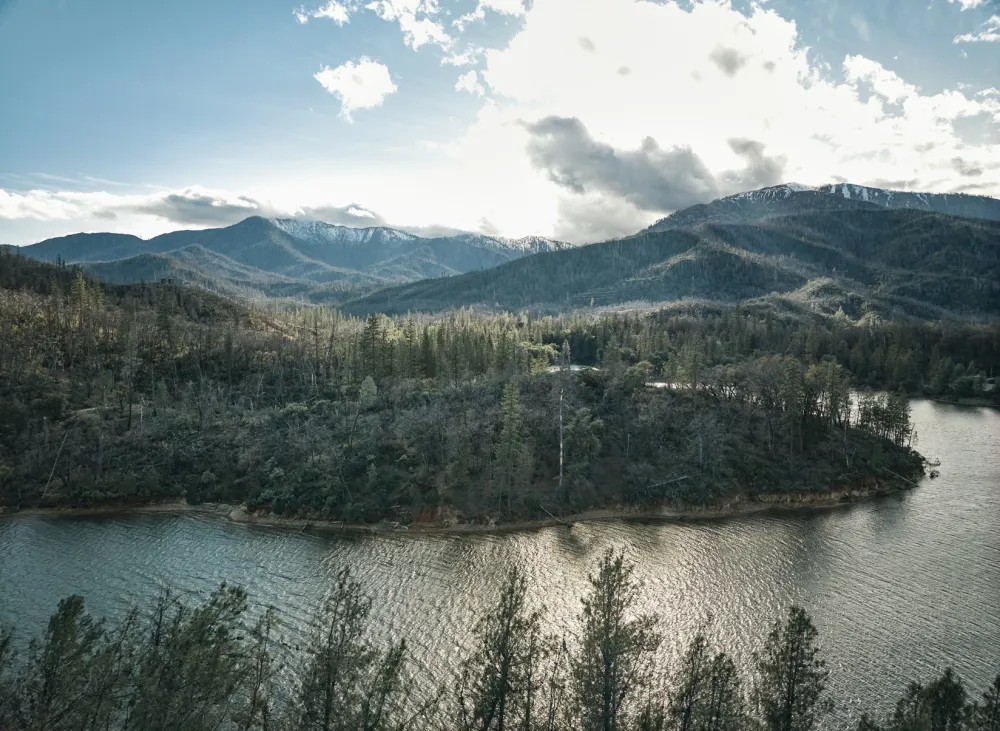 Lush green hills and a serene river reflect sunlight under a cloudy sky, framed by distant snow-capped mountains.