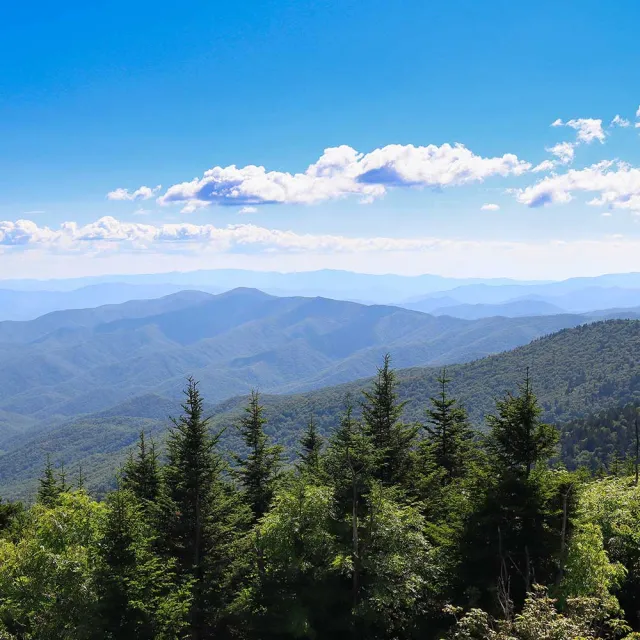 Vast blue sky over layered mountain ranges, dotted with green trees, creating a serene landscape of nature's beauty.