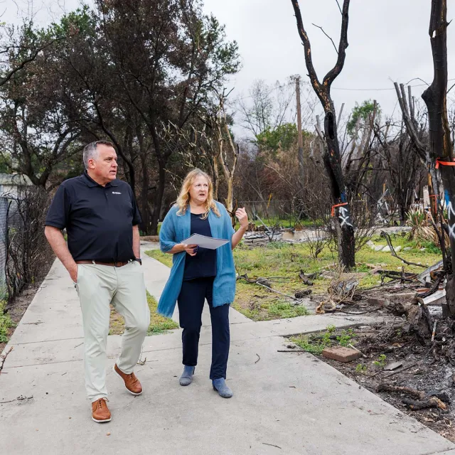 A woman and a man discuss plans while walking through a landscape affected by recent wildfire damage, marked by charred trees and debris.