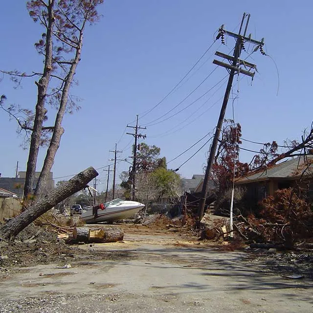 Devastated street with uprooted trees, debris, and a stranded boat, reflecting the aftermath of a natural disaster under a clear sky.