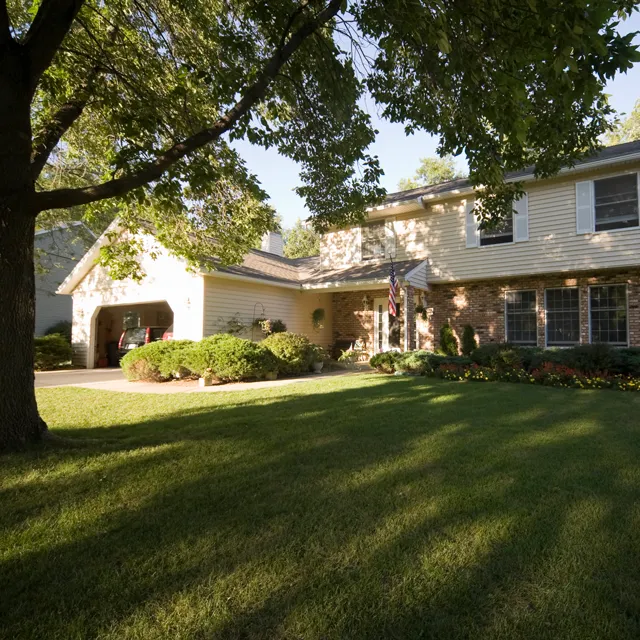 A two-story suburban home surrounded by green grass and trees, featuring a porch and an American flag hanging by the entrance.