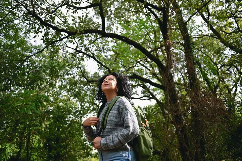 A person stands in a lush green forest, wearing a cozy sweater and a backpack, enjoying the peaceful outdoor surroundings.
