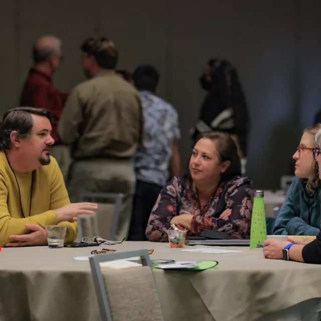 People talking with each other around a table, with several others standing in the background.