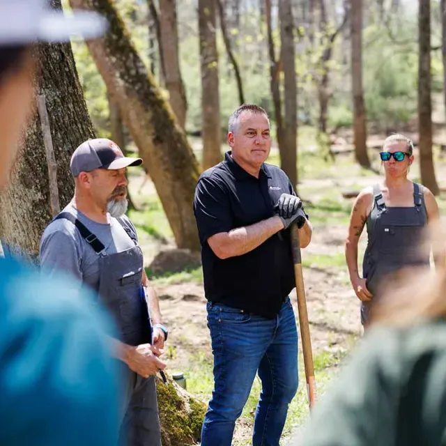 A group of people stands outdoors in a forested area, discussing plans while holding gardening tools and wearing work clothes.