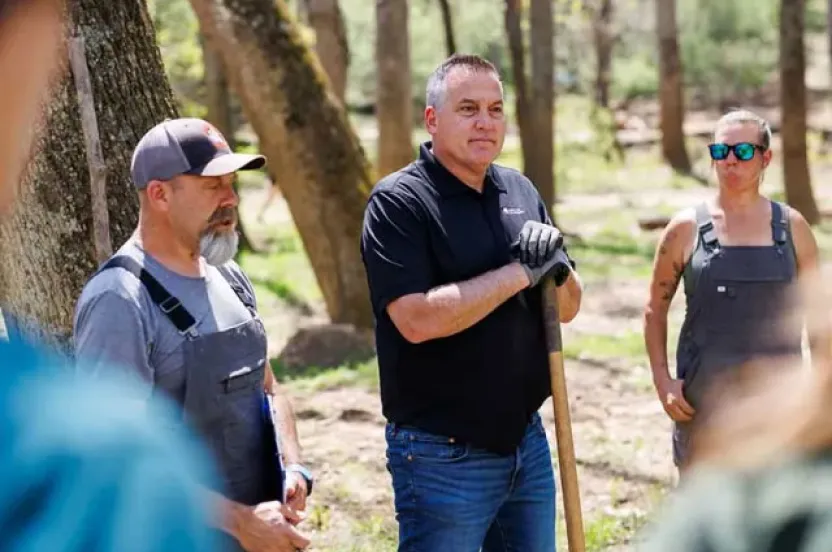 A group of people stands outdoors in a forested area, discussing plans while holding gardening tools and wearing work clothes.