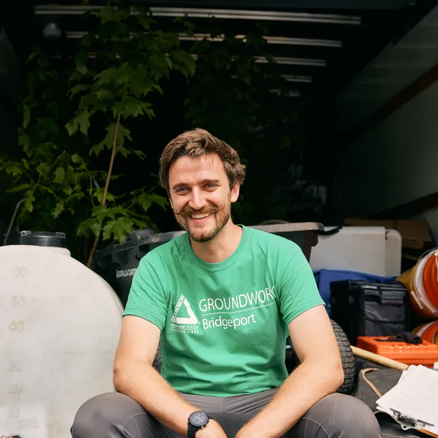 A person in a green Groundwork Bridgeport t-shirt sits among gardening tools and plants in a truck, ready for work.