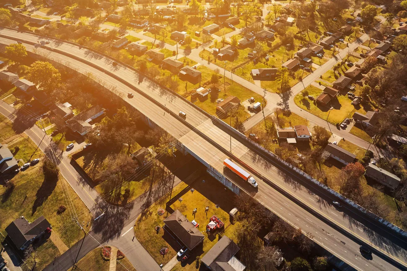 Aerial view of a suburban neighborhood with houses, streets, and a busy highway featuring a truck and cars against a sunset backdrop.