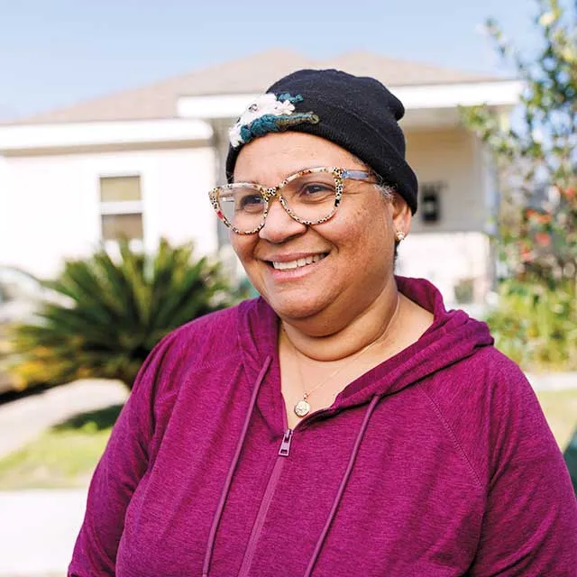 A person in a purple zip-up hoodie and glasses stands outdoors with a house and lush plants in the background on a sunny day.