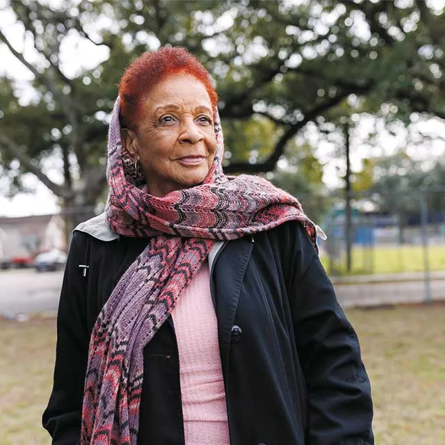 A woman with short, dyed red hair wears a patterned scarf and a black jacket, standing outdoors with trees in the background.