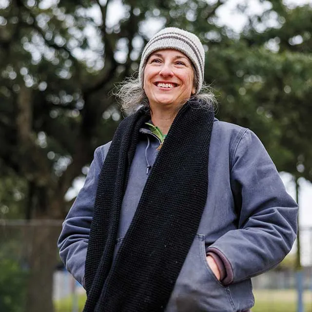 A woman wearing a striped beanie, navy jacket, and a long black scarf stands outdoors, surrounded by leafy trees.