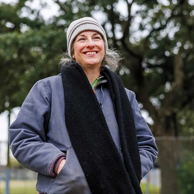 A woman with long gray hair, wearing a gray jacket, black scarf, and a striped beanie stands outdoors against a leafy backdrop.