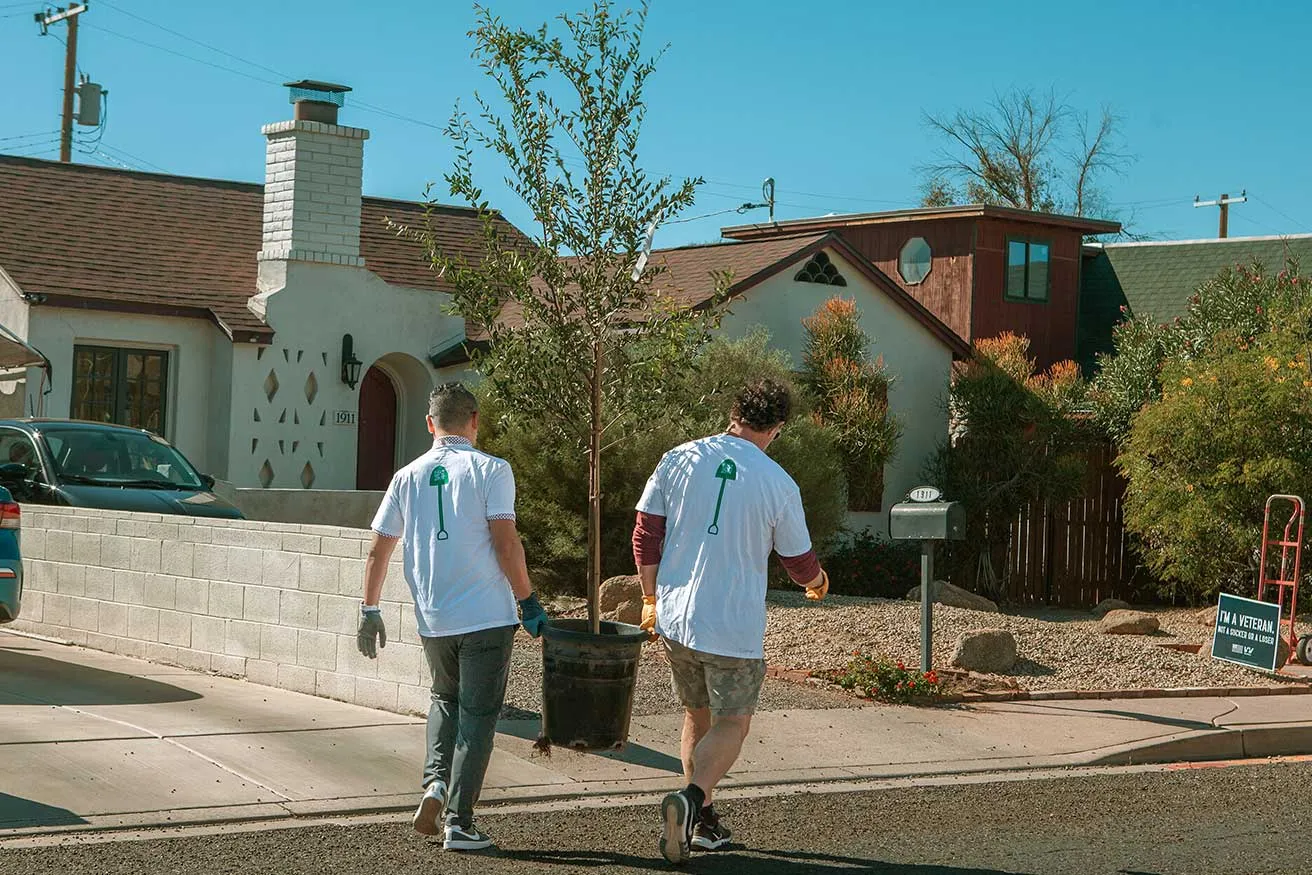 Two volunteers in matching shirts carry a potted tree down a residential street, ready to plant it in a sunny neighborhood.