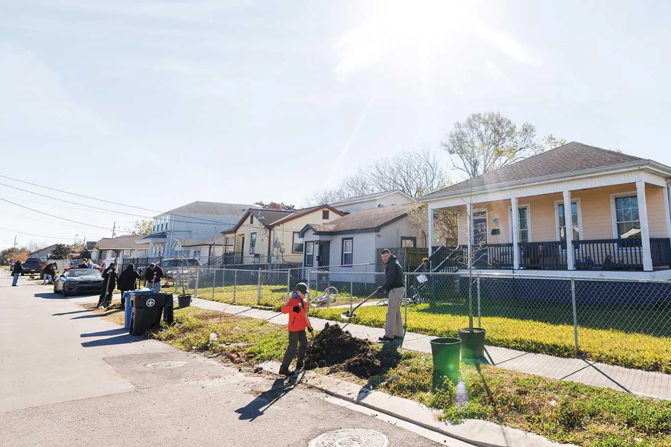 A man and a boy dig a hole in a sunny suburban street, surrounded by neighbors cleaning up yards and parked cars.