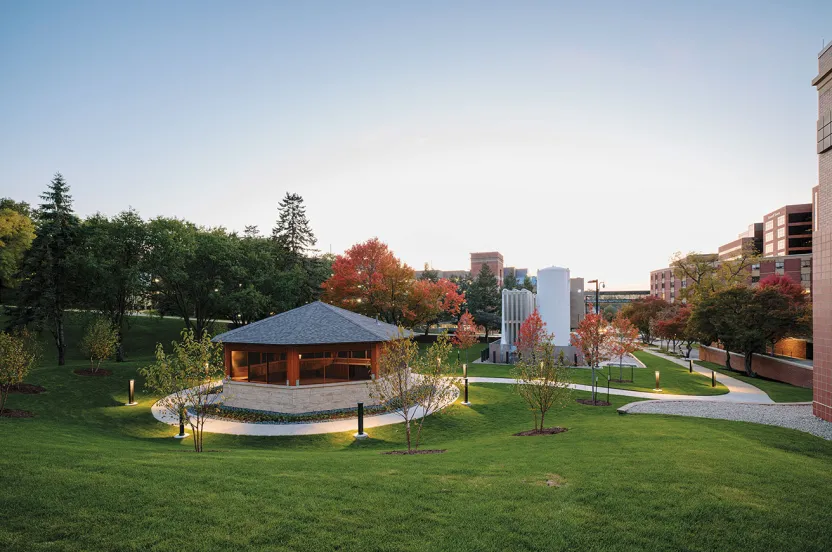 A landscaped courtyard features a wooden gazebo, colorful autumn trees, and modern sculptures, all under a clear blue sky.