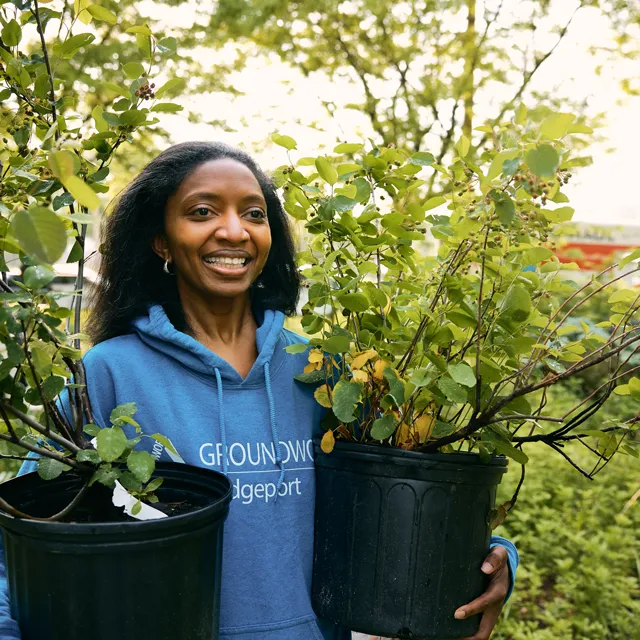 A person in a blue hoodie is holding two large pots filled with green plants, surrounded by greenery in a bright outdoor setting.