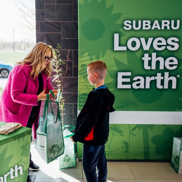 A child receives a reusable bag filled with eco-friendly items from a woman, in front of a Subaru promotional banner about environmental care.