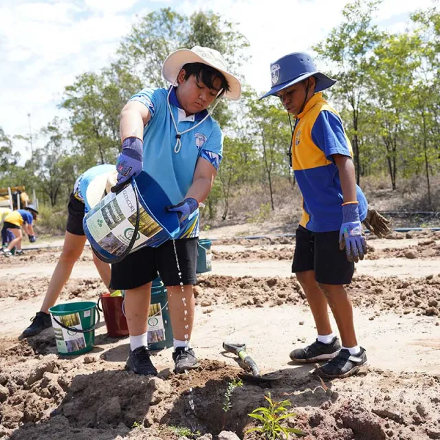 Two children in hats pour water from a bucket into a hole in the ground, surrounded by planting materials in a sunny outdoor setting.
