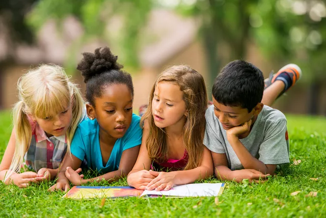4 children laying in grass reading something together.