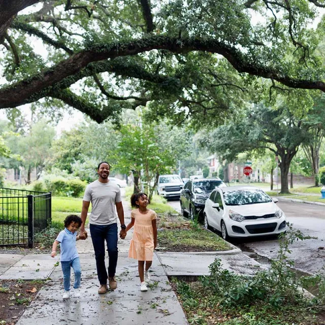 A parent walks hand-in-hand with two children along a tree-lined street, surrounded by greenery and parked cars.