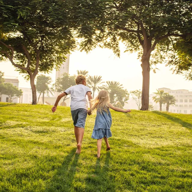 Two children, a boy and a girl, run hand-in-hand across a sunny green field, surrounded by trees and a glowing sunset.