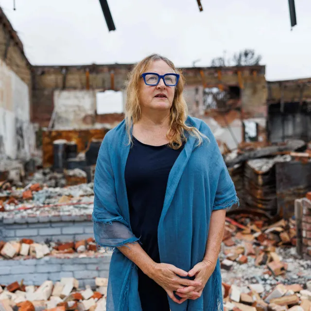 A woman in a blue shawl stands amidst the ruins of a building, surrounded by scattered bricks and debris under a cloudy sky.
