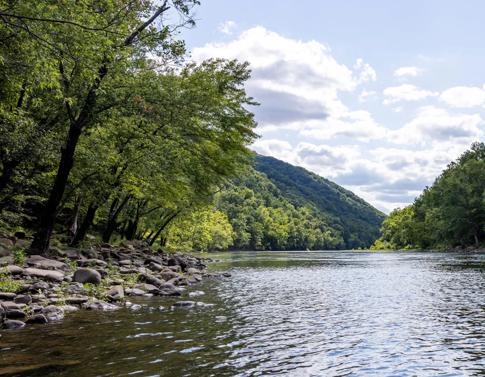 Serene river scene with gentle waters reflecting clouds, surrounded by lush green trees and rolling hills under a bright sky.