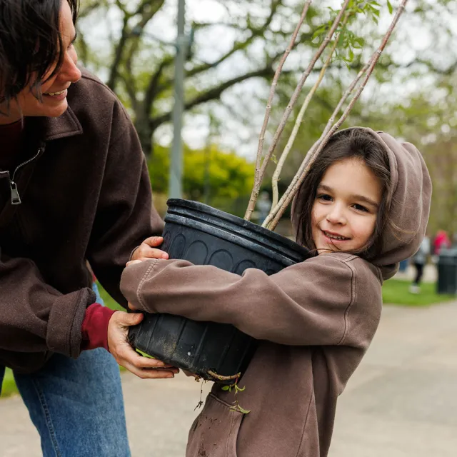 A child in a brown hoodie hugs a large plant pot while an adult assists them, surrounded by greenery in a park setting.