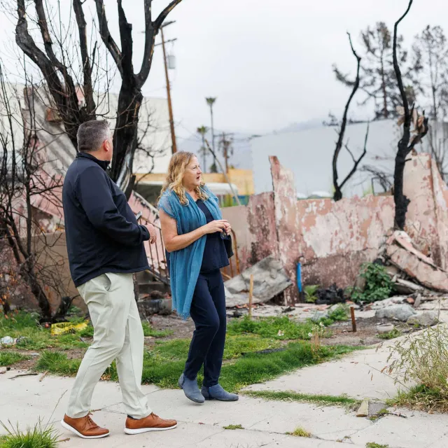 A man and woman walk through a neighborhood with fire-damaged buildings and charred trees. The mood appears somber as they assess the destruction.