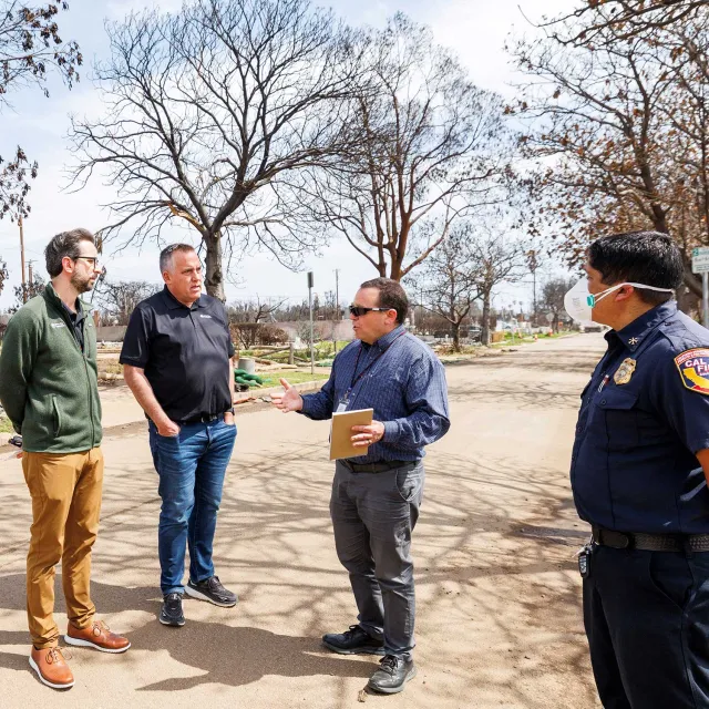 Four men stand in discussion on a barren street surrounded by leafless trees. One wears a uniform and mask, conveying a serious tone.